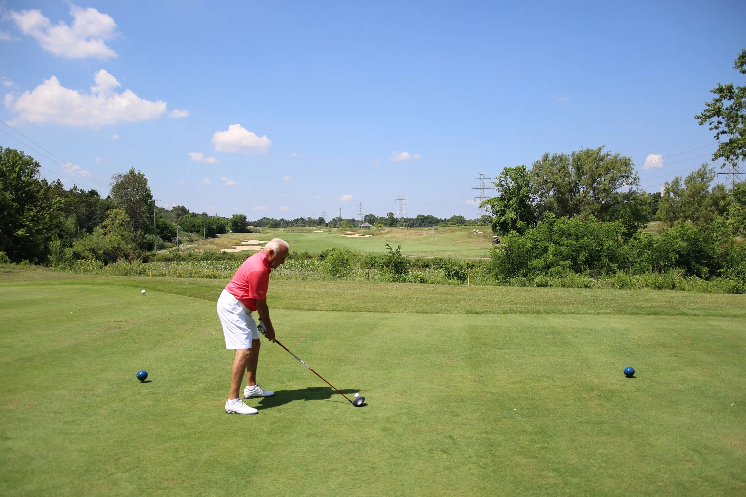 A man wearing a pink shirt and white shorts playing golf on a grassy course under a blue sky with some clouds.