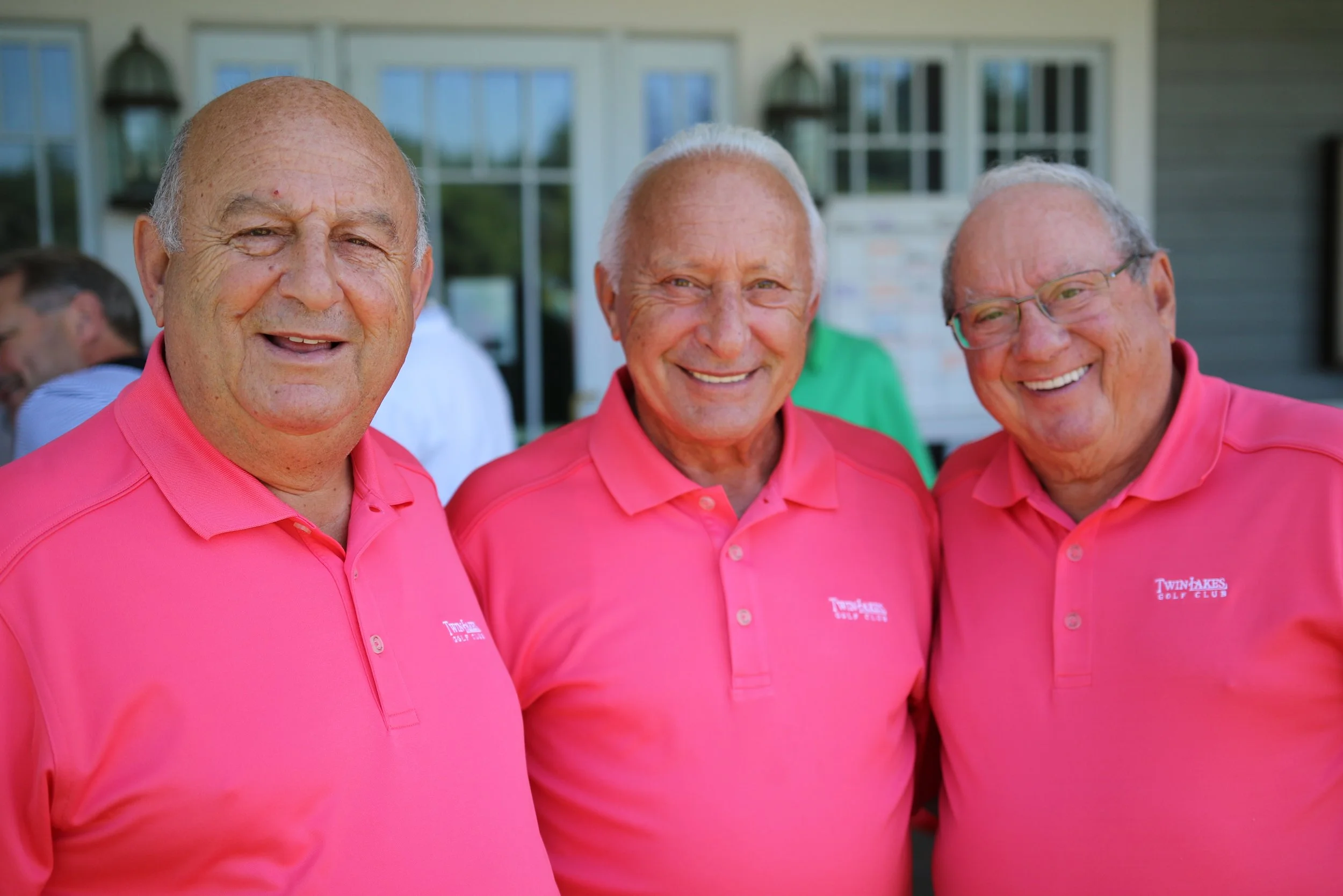 Three elderly men wearing matching pink polo shirts smiling outdoors.