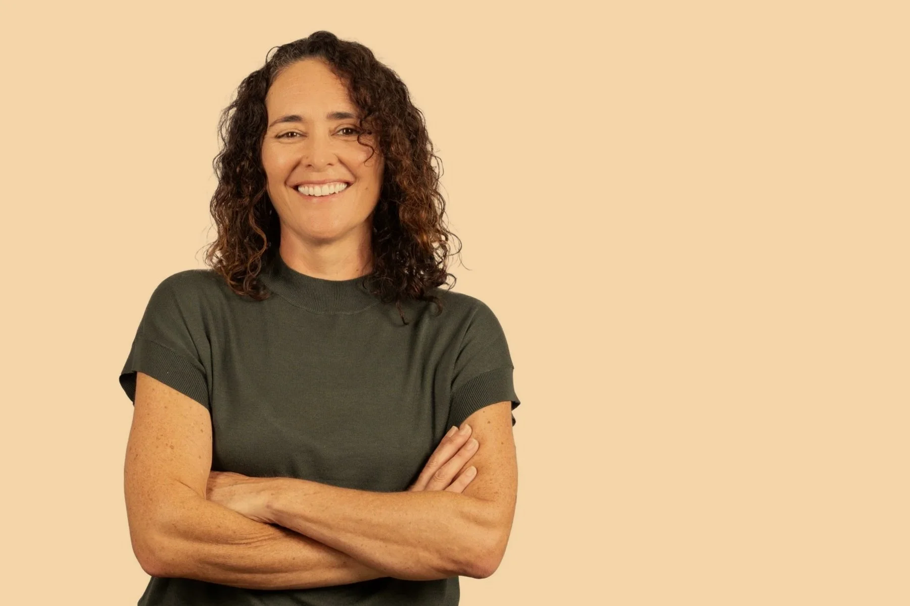 A woman with curly brown hair smiling, wearing a dark grey t-shirt, standing against a beige background with her arms crossed.