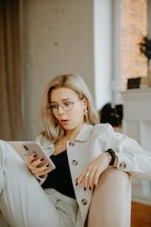 Young woman with glasses looking at her phone while sitting on a couch in a well-lit room.