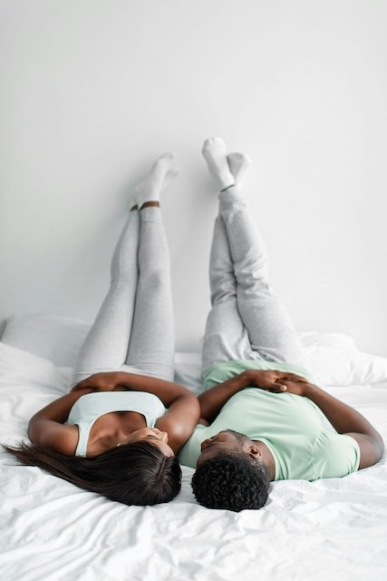 A woman and a man lying on a bed with their legs extended upward against a white wall, relaxing.