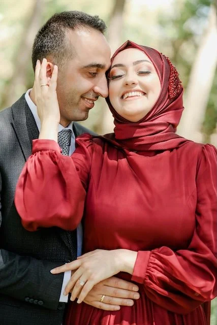A couple is smiling and touching foreheads outdoors, the woman wearing a red dress and hijab, the man in a suit.
