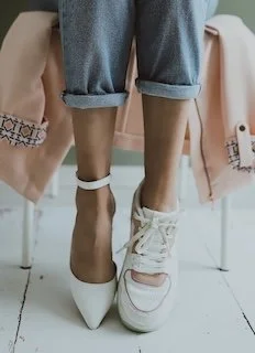 Close-up of a person's lower legs wearing rolled-up jeans and mismatched shoes: a pointed white heel on one foot and a casual white sneaker on the other, with a pink coat draped over a chair in the background.