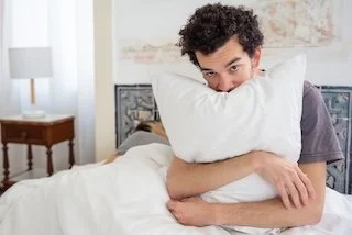 A man hugging a pillow in bed, looking at the camera.