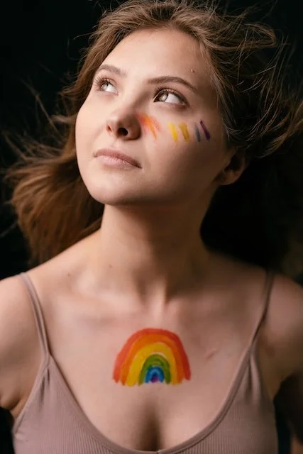 A young woman with rainbow face paint on her cheek and a rainbow painted on her chest, looking upward with a neutral expression.