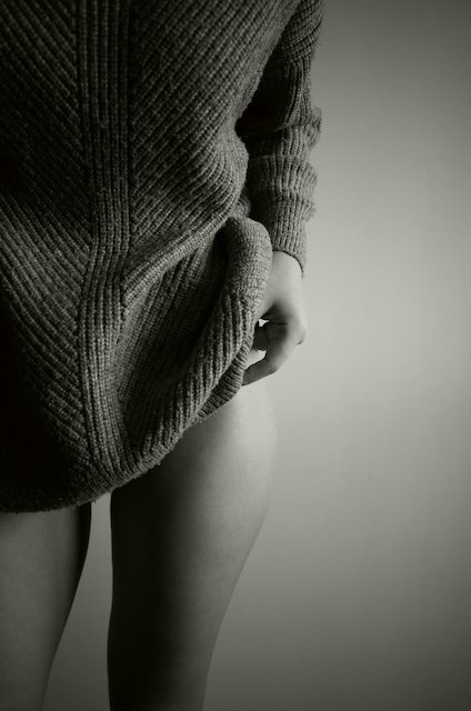 Close-up of a person in a knitted sweater, holding the sweater's fabric near their thigh, with a plain background.