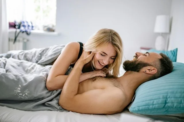 A woman and a man are in bed, smiling and looking at each other in a bright bedroom.