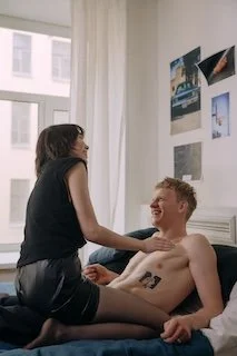 A woman with glasses and dark hair standing and interacting with a smiling shirtless man lying in bed, in a well-lit room with large windows and framed pictures on the wall.