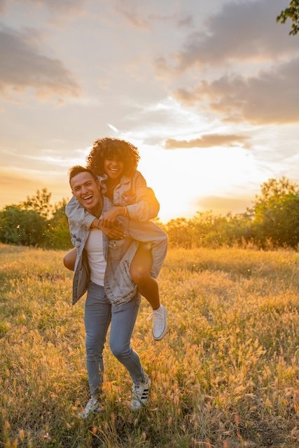 A happy young woman with curly hair piggybacking on a smiling young man in a grassy field at sunset.