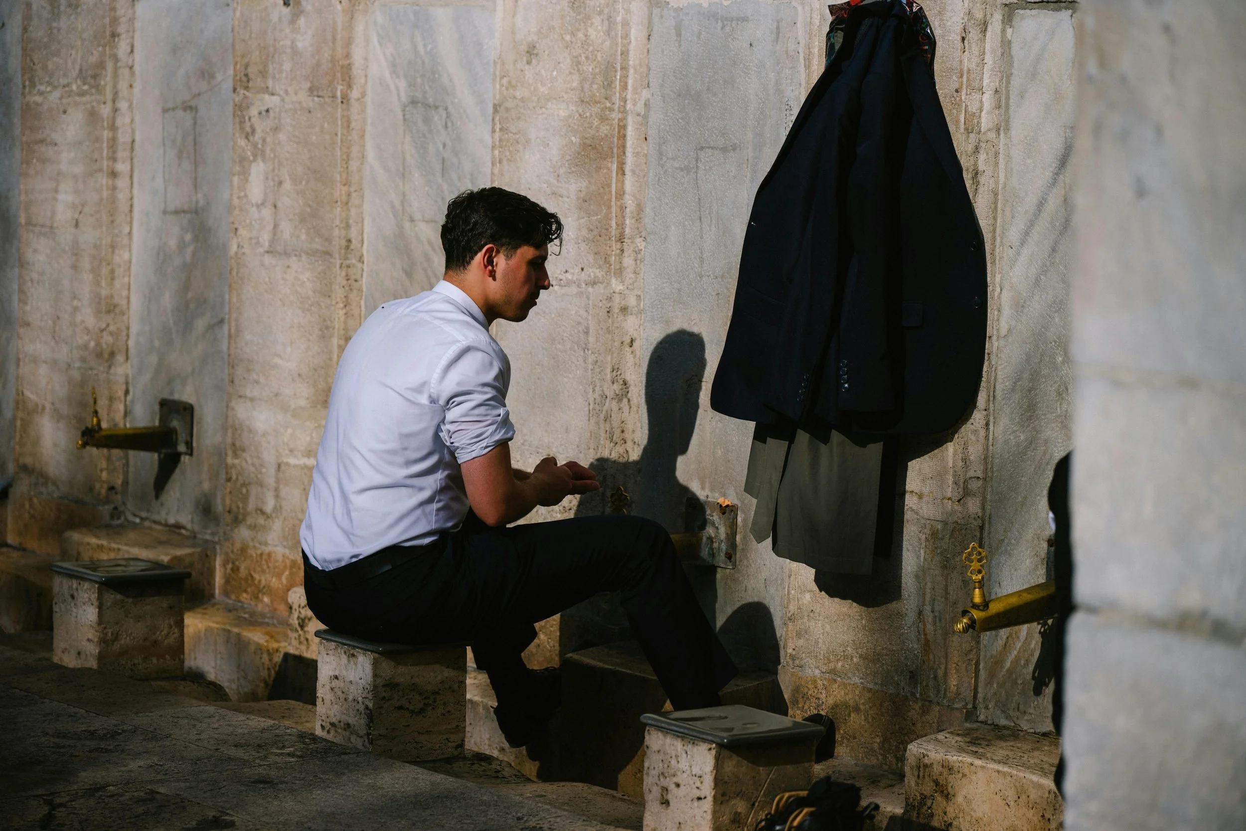 A young man with dark hair, dressed in a white shirt and black pants, kneels on a stone bench in front of a stone wall with a water fountain attachment, waiting to wash his face or hands, with a black jacket hanging on the wall behind him.