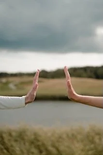 Two hands held up with palms facing each other, over a natural landscape with a body of water and cloudy sky.