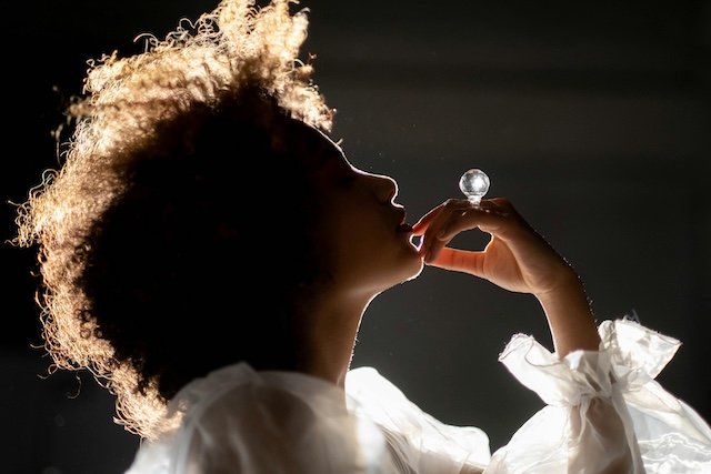 Silhouette of a woman with curly hair holding a glass orb near her lips, backlit to create a glowing outline, wearing a white, ruffled garment.