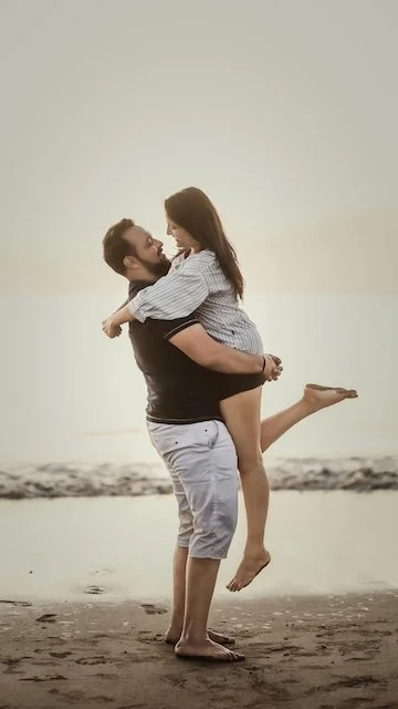 A man lifting a woman on a beach at sunset, both smiling and embracing.