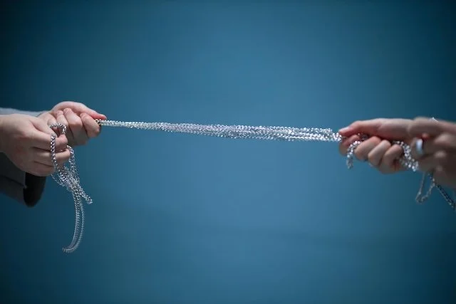 Two hands pulling a long string of shiny silver beads or jewelry against a plain blue background.