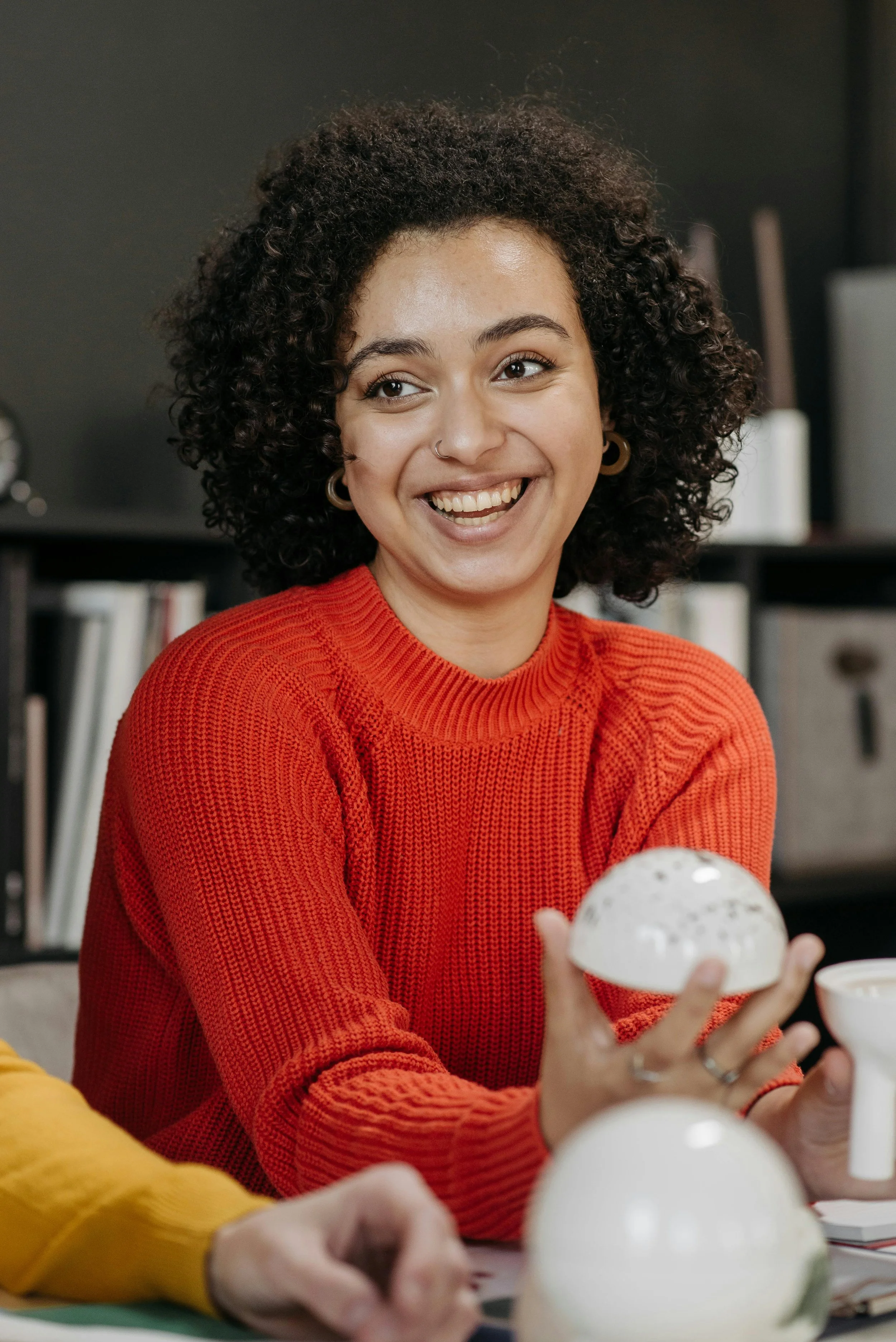 A woman with curly hair, wearing a red sweater and hoop earrings, smiling and holding a white ceramic bowl in her right hand, sitting at a table with others. The background has a dark wall and bookshelves.