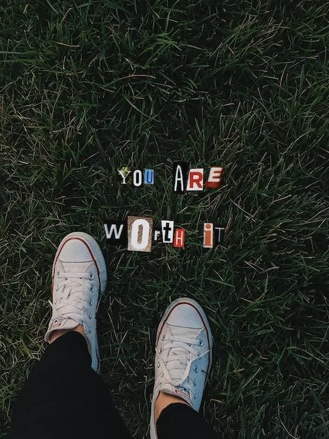 View of white sneakers and colorful cut-out letters on grass spelling 'YOU ARE WORTH IT'.
