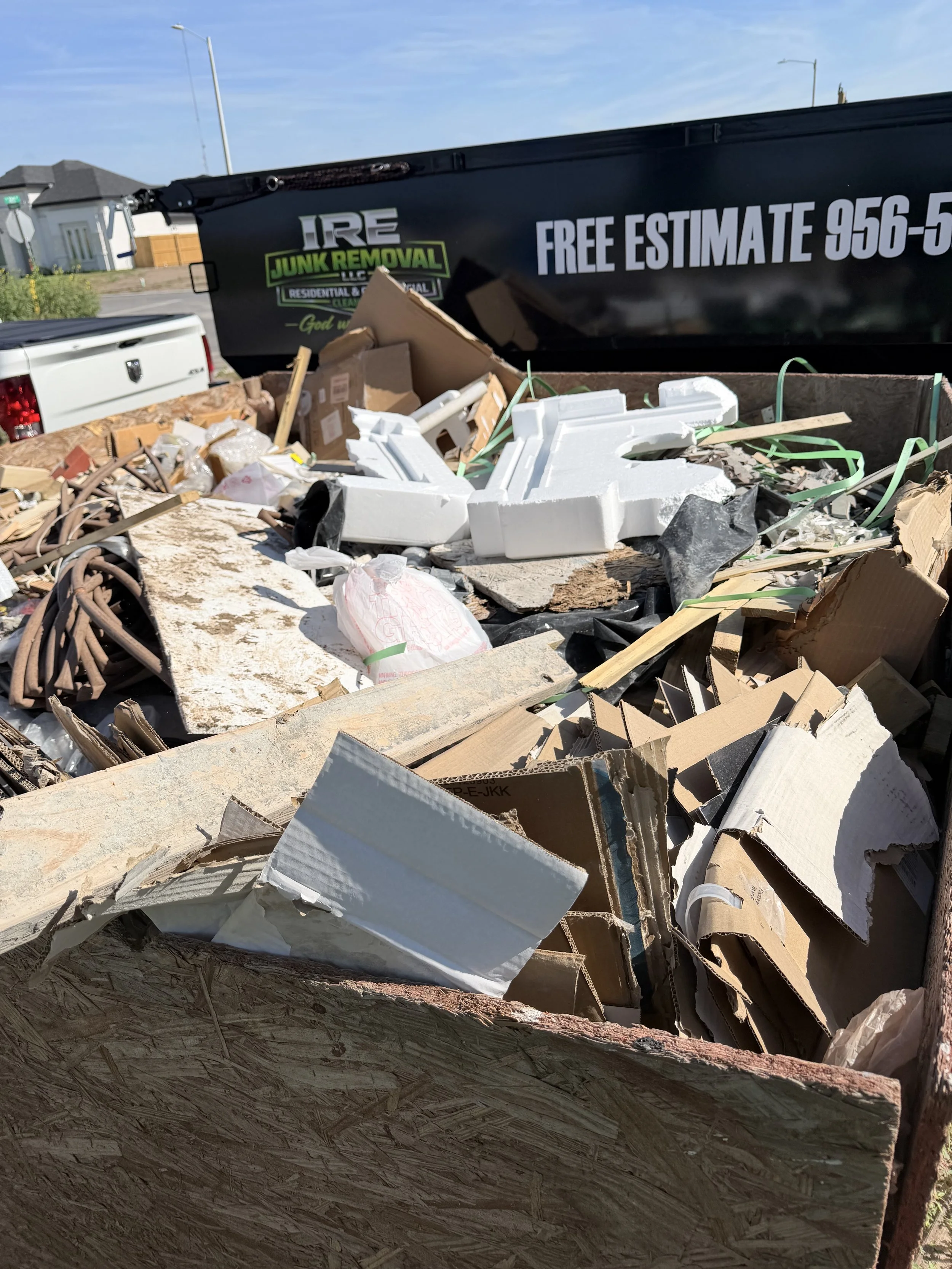 A large black dumpster filled with debris, including cardboard, foam, and wood, with a white pickup truck nearby on a residential street under a partly cloudy sky.