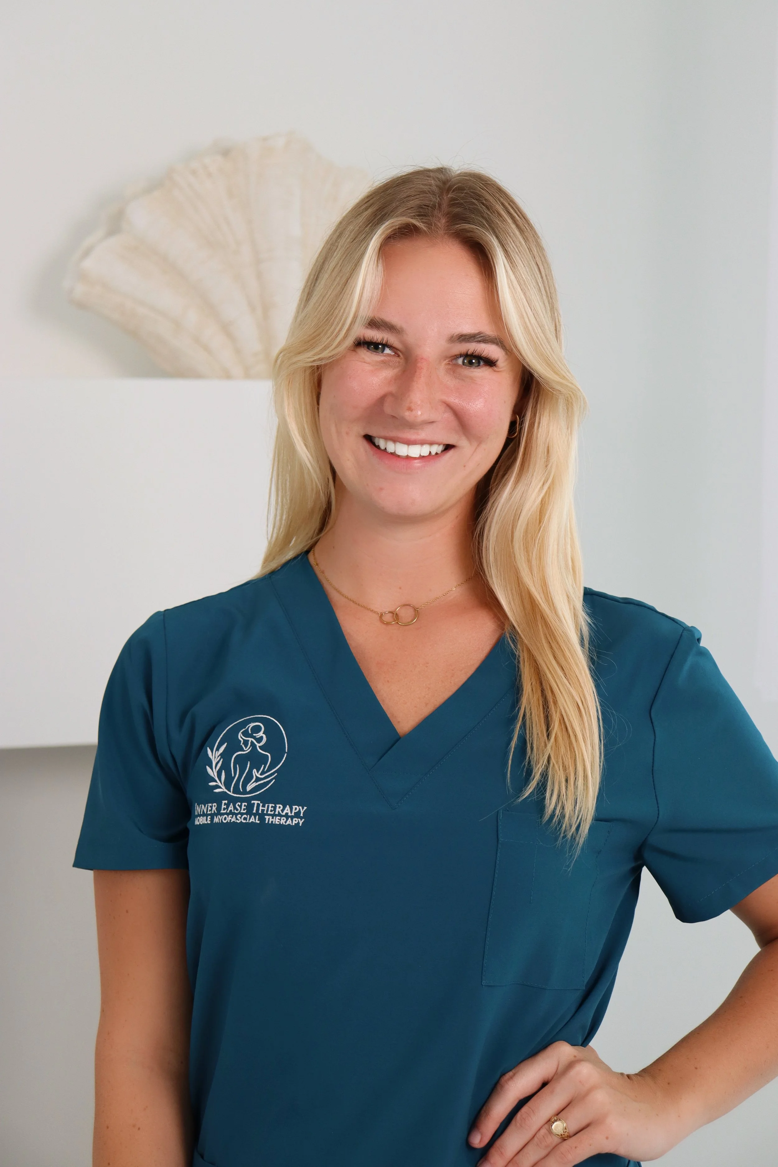 Woman in teal medical scrubs smiling, standing against white background, with seashell decoration on the wall behind her.