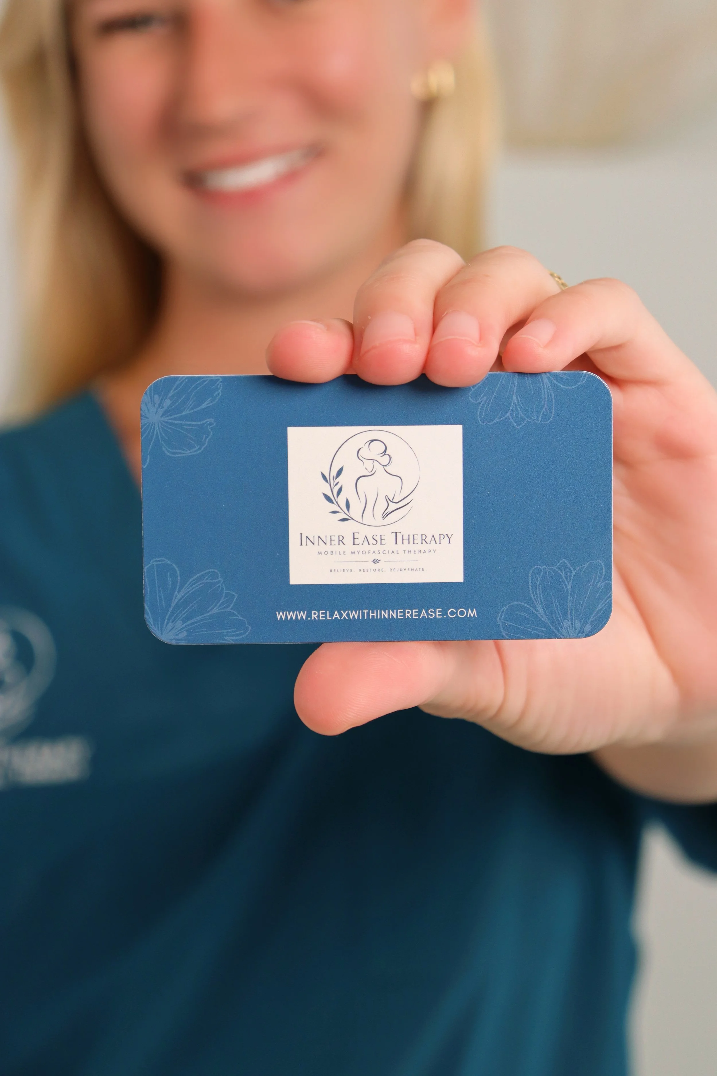 A woman with blonde hair smiling and holding a blue card with the logo and text for Inner Ease Therapy, a mobile myofascial therapy service.