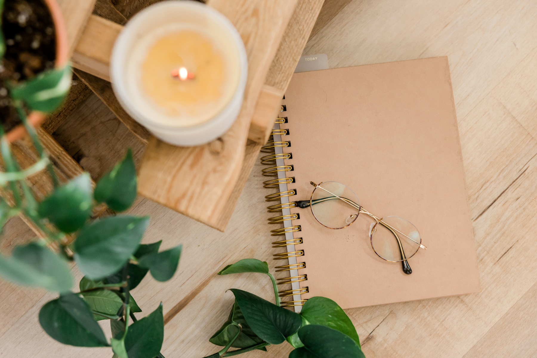 A flat lay of a desk with a candle, a potted plant, a spiral notebook, glasses, and a brown paper notebook on a wooden surface.