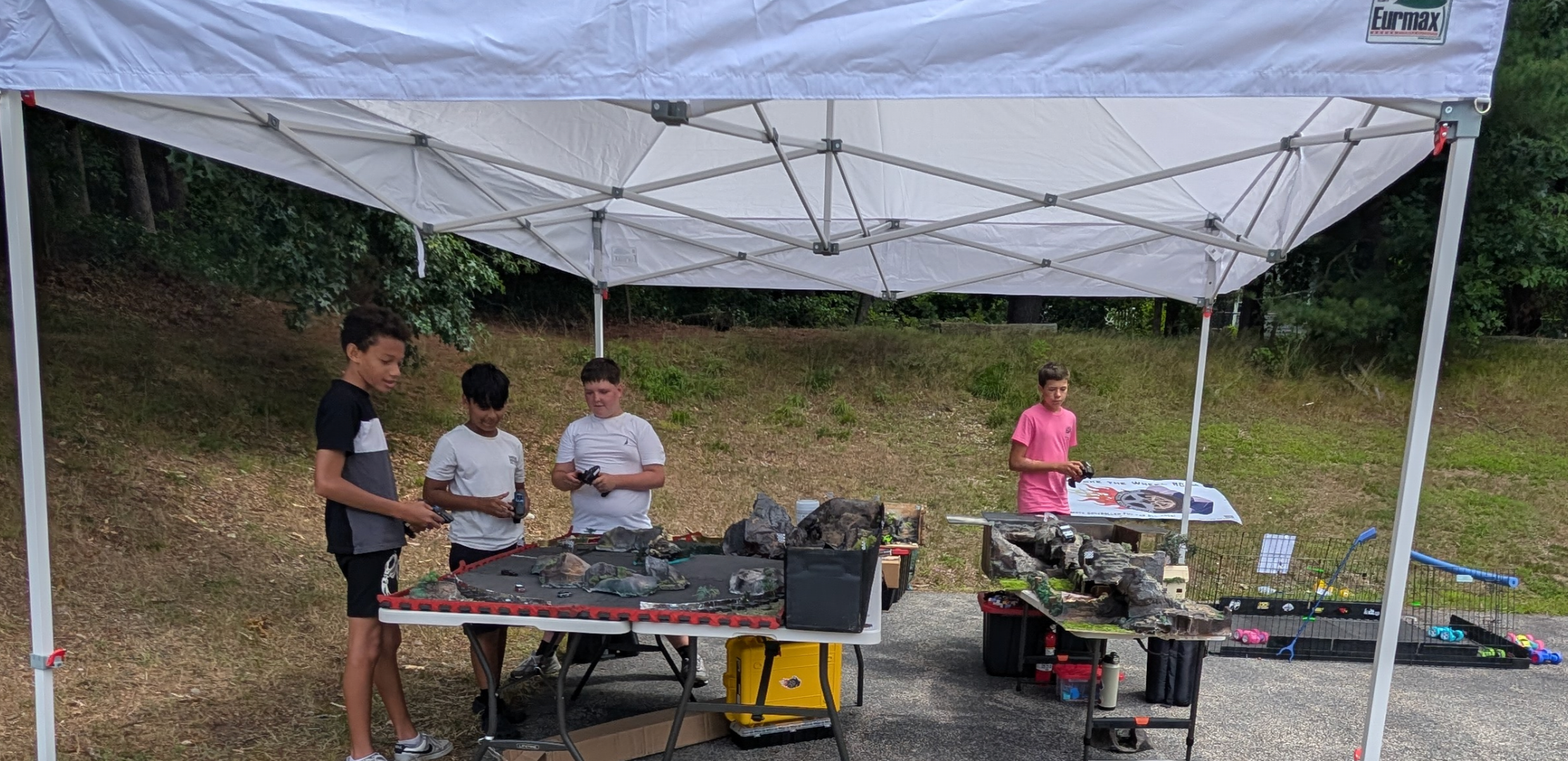 Four boys standing under a white canopy tent at an outdoor event, with tables displaying rocks and tools, on a grassy hillside.