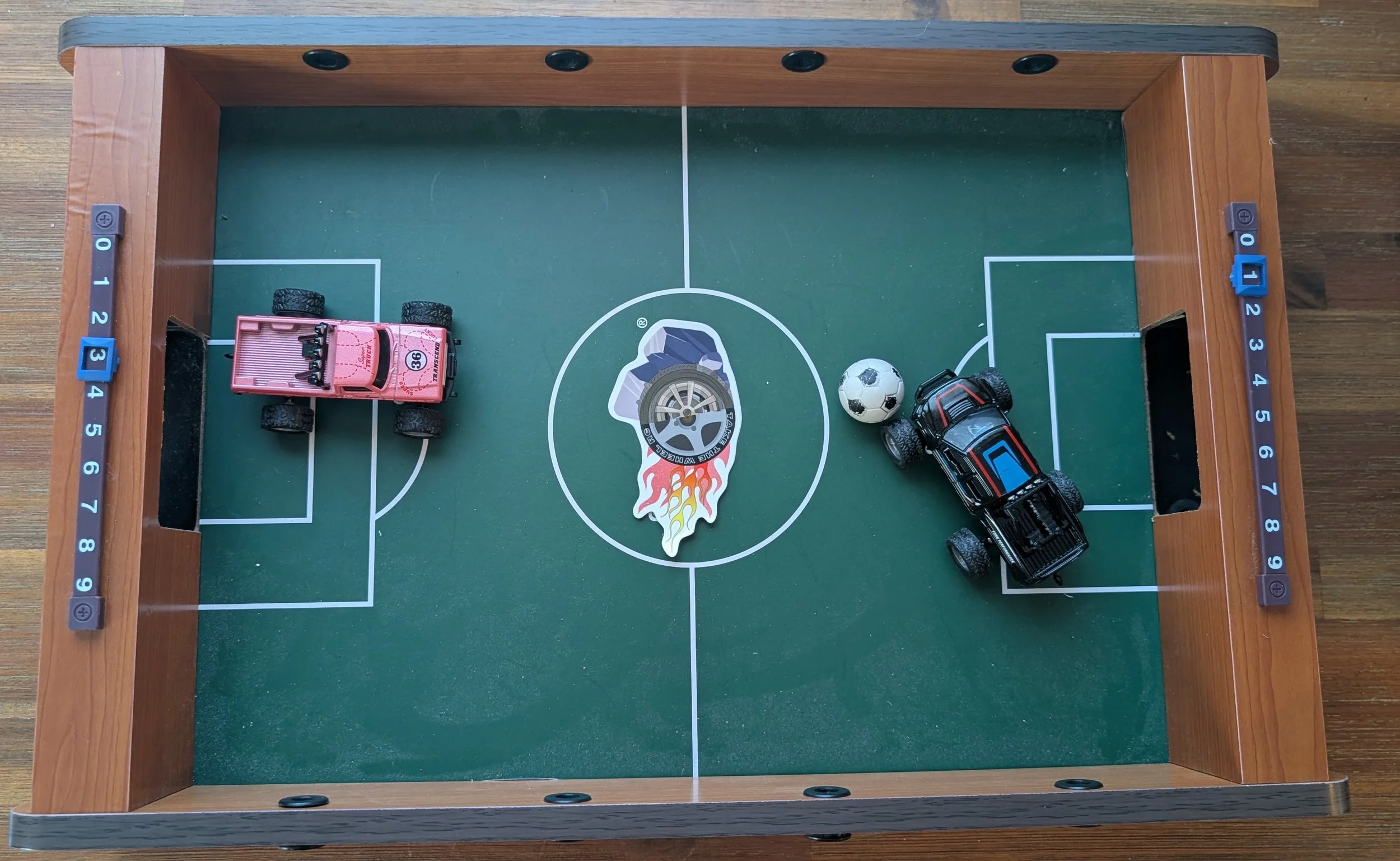 A tabletop soccer match featuring a mini soccer field with two micro RC trucks positioned as players, a soccer ball, and score markers on the sides. The field has a central Take The Wheel RC logo