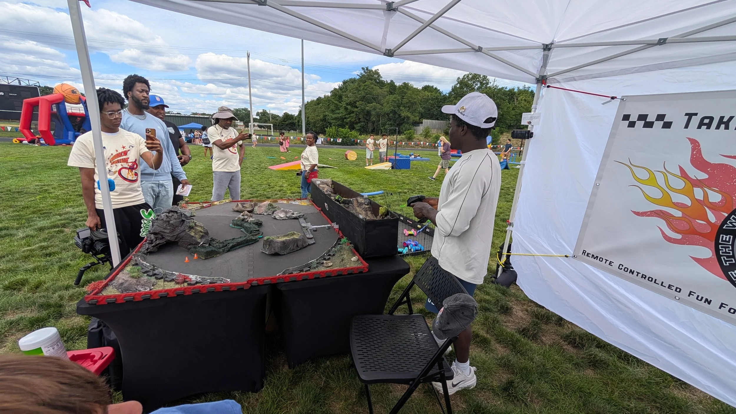FredFlips takes the wheel at the Frederick Flips Richard fair in Stoughton,  with large model terrain for a remote-controlled car race, under a white canopy tent on a grassy field, 