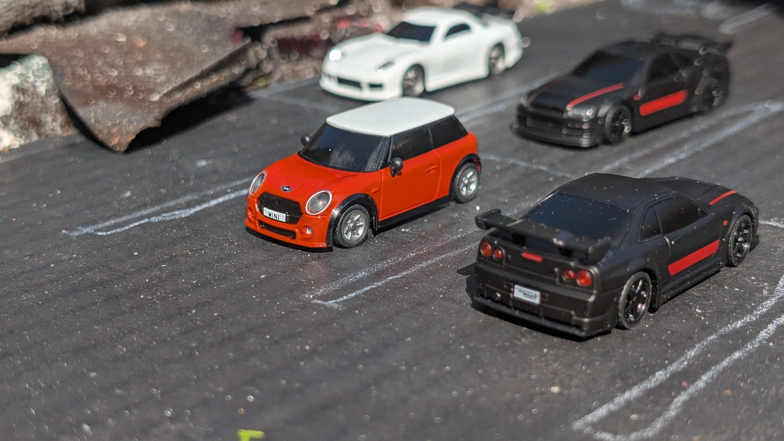 Close-up of miniature race cars parked on a miniature parking lot, including a red and black Mini Cooper, a white sports car, and two black sports cars with red accents.