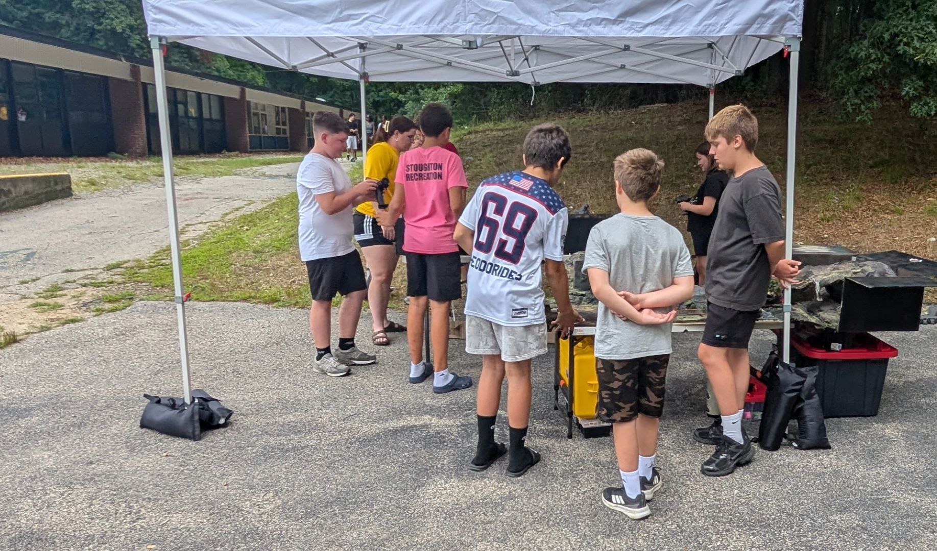 A group of children and teenagers gathered under a white canopy tent outdoors, around multiple race track tables, with a school building and trees in the background.