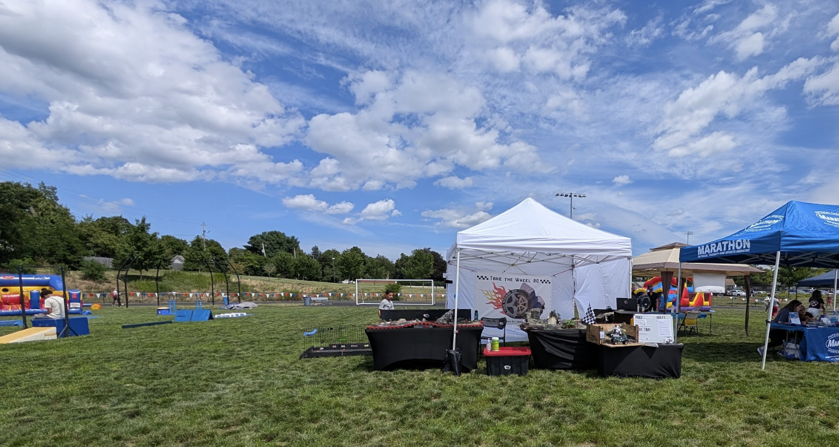 Outdoor community event on a grassy field with tents, tables, and inflatable bounce houses, under a partly cloudy sky.
