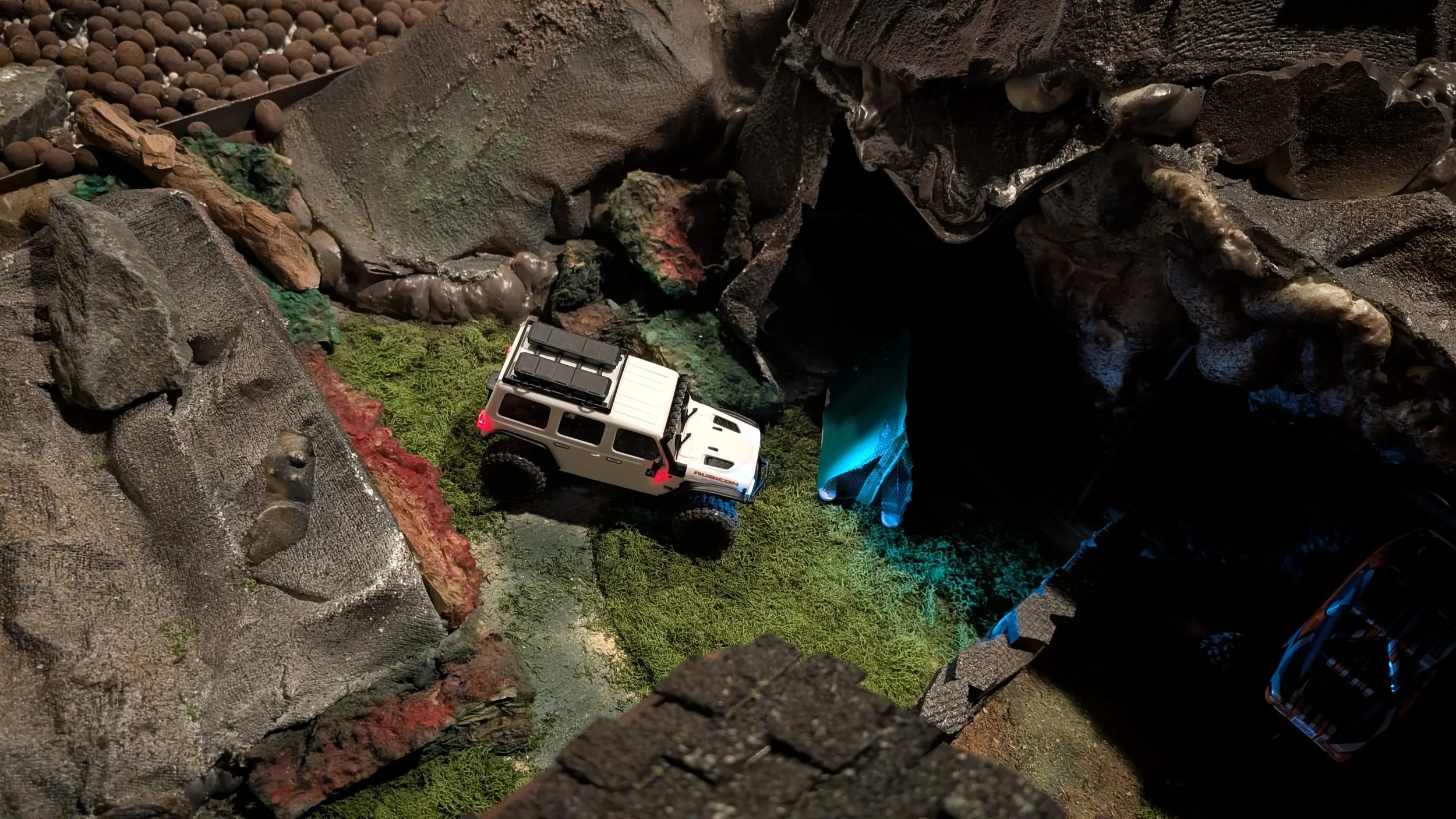 Model of a white Axial Jeep Wrangler Rubicon SCX30, off-road vehicle on a miniature terrain with rocks, moss, and a small campers tent.