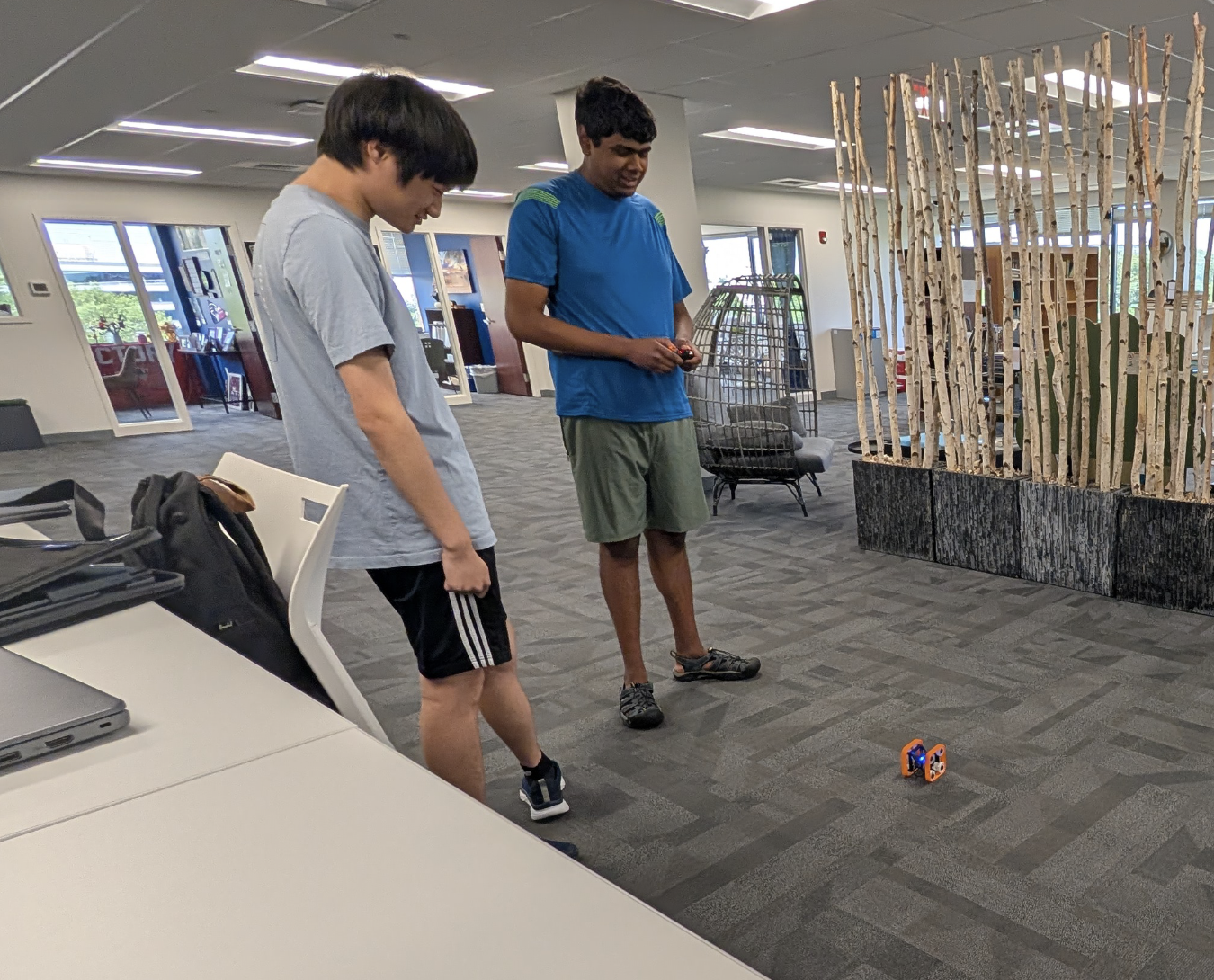 Two young men watch a small remote-controlled robot they built in a STEM robotics workshop on the carpeted floor of an office.