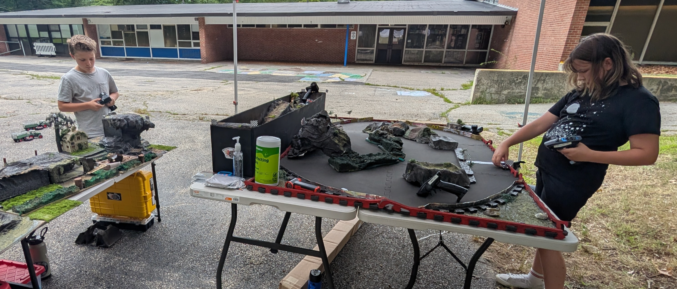 Two children driving cars on the race track and rock crawler track set outdoors in front of a school.