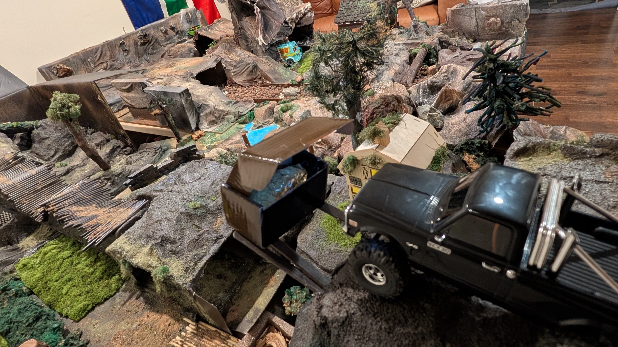 A detailed miniature model scene featuring a black toy pickup truck on a rocky terrain with small trees, shrubs, and miniature vehicles.
