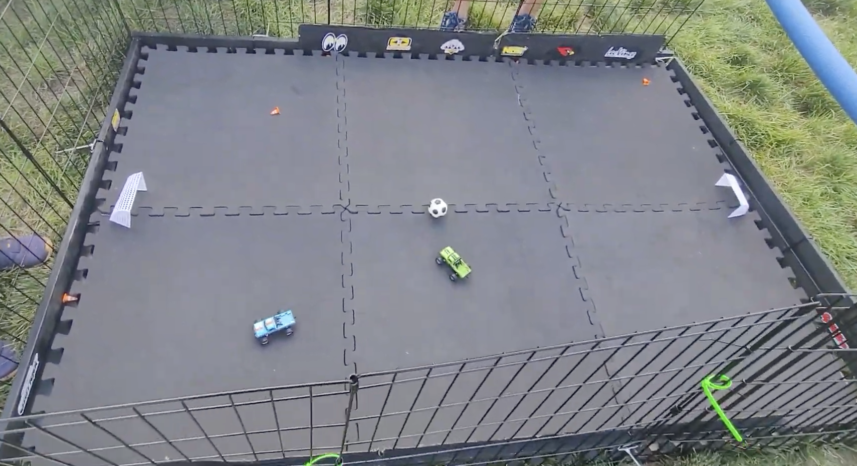 A small soccer or hockey game set up inside a fenced outdoor area. There are toy cars on the court, with a small white ball, two goals, and some small accessories, including icons on the fence.