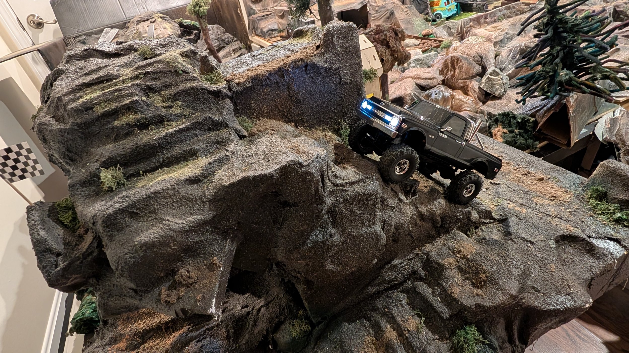 A detailed model of a rocky terrain with small plants and a black remote-controlled off-road truck climbing the rocks.