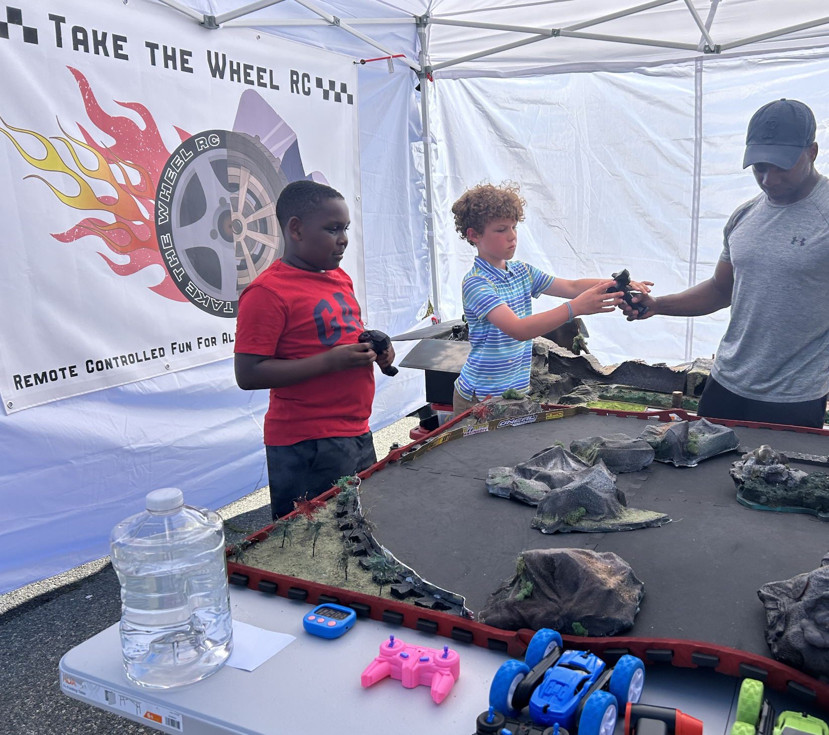 An adult hands over an RC transmitter to a participant while cars are racing on a track in a tent at an outdoor event. The tent has a sign that reads 'Take the Wheel RC' and features a large race car illustration with flames.
