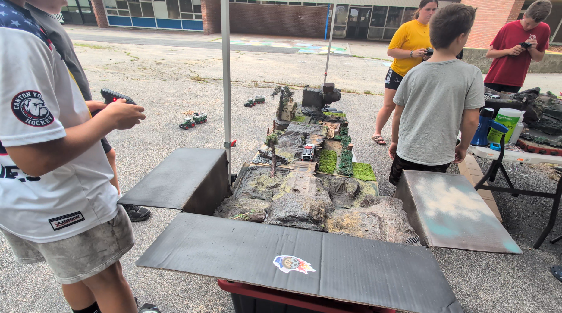 Children and teenagers gathered around a tabletop diorama of a miniature landscape with small toy vehicles, trees, and rocks, part of a model train or war game setup outdoors.