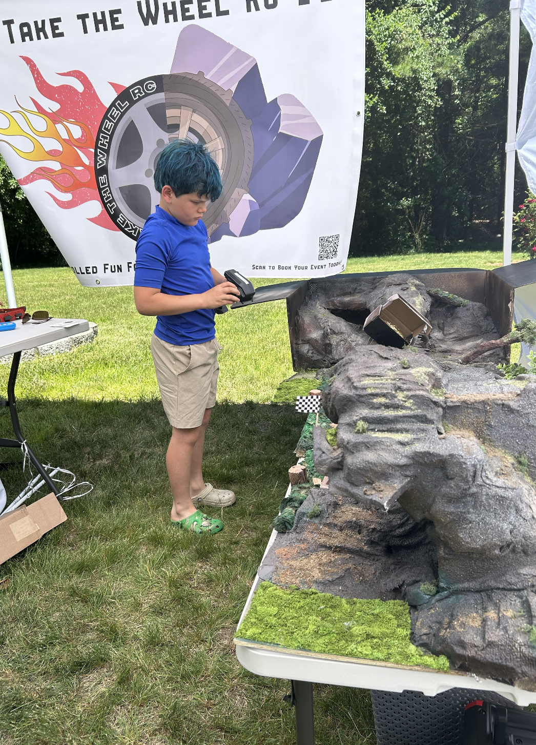 A boy with blue dyed hair using an RC crawler at an outdoor birthday party. He stands next to a rocky artificial landscape with a small RC Ford Bronco on a slope. 