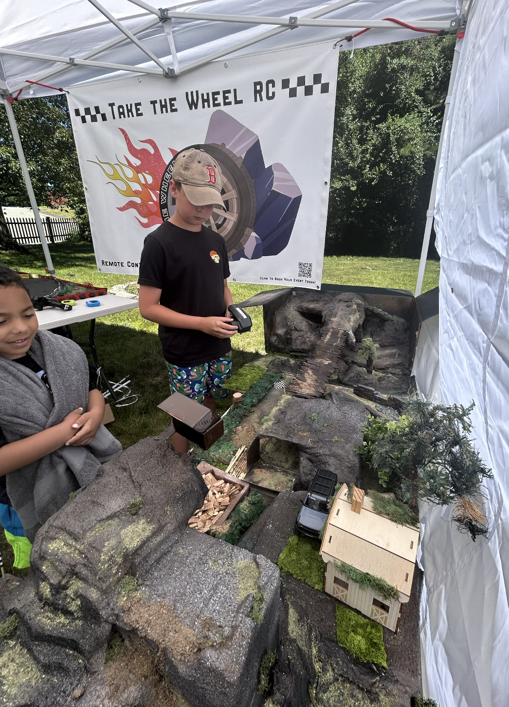 Children at a RC car racing event, with a model racetrack set up inside a tent, featuring miniature houses, trees, and a ramp for RC cars, and a banner that reads 'Take the Wheel RC' behind them.