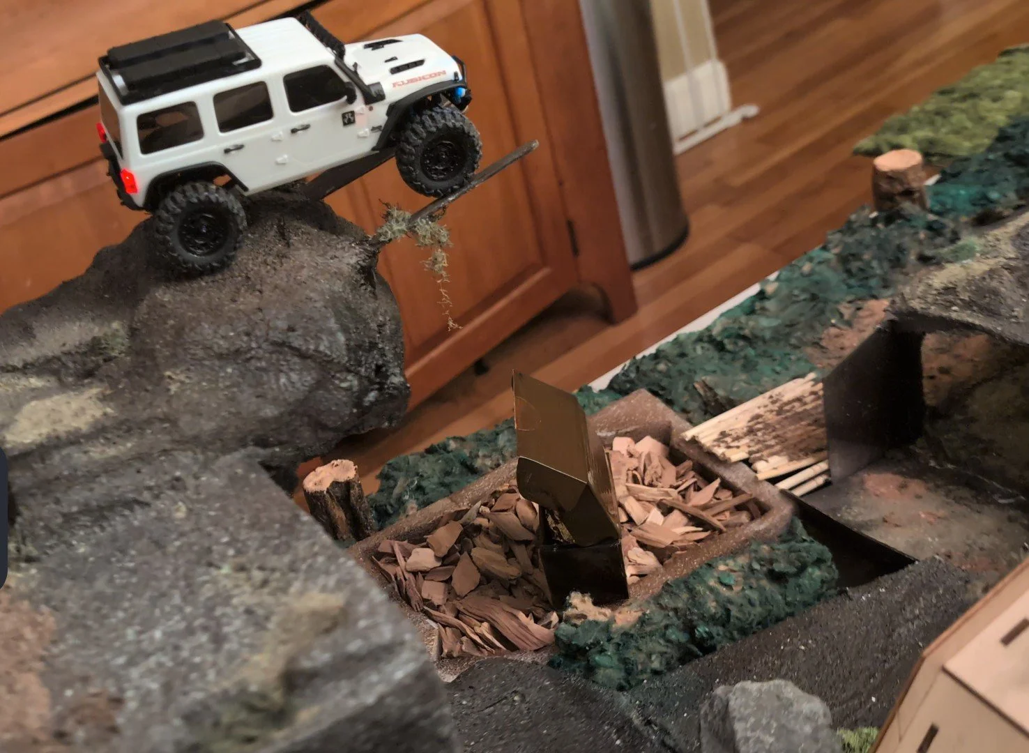 Axial SCX30 Jeep Wrangler Rubicon positioned on a summit, appearing to climb a steep incline. The background features indoor furniture, including wooden cabinets and a floor grate or vent, indicating that the scene is set at a indoor event.