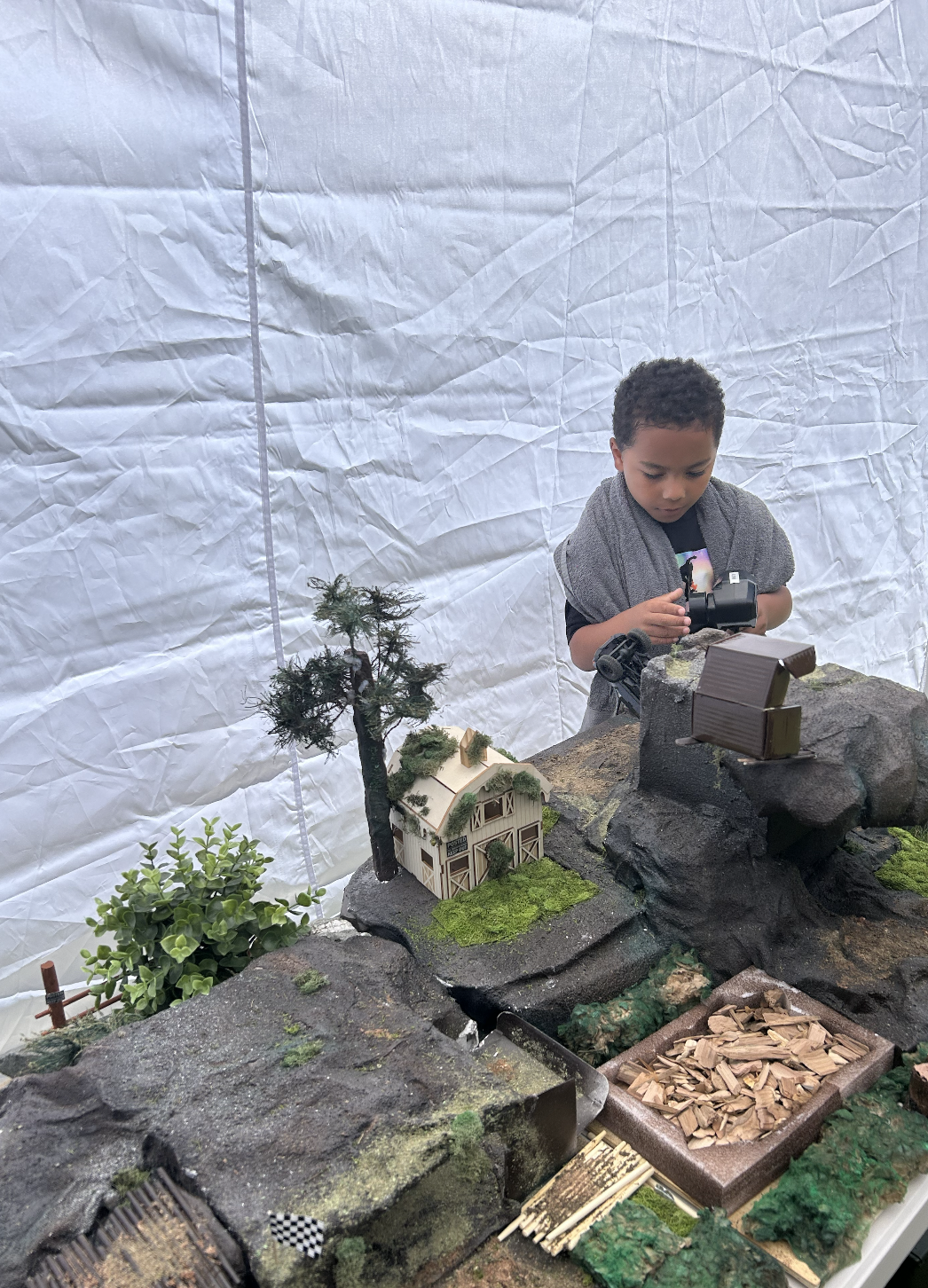 A young boy standing behind a miniature model landscape, holding an RC transmitter, inside a tent at an event.