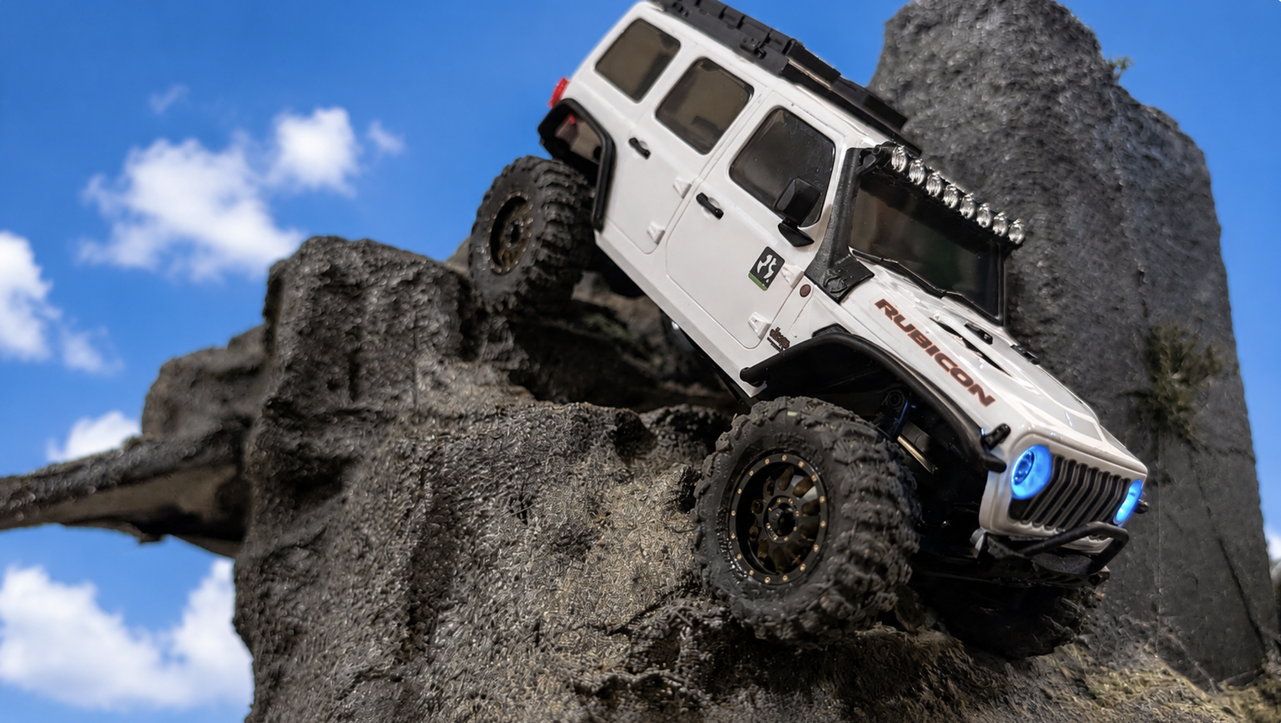 A white remote-controlled off-road vehicle navigating rocky terrain with large tires, blue LED lights, and a rugged design against a bright blue sky with white clouds.
