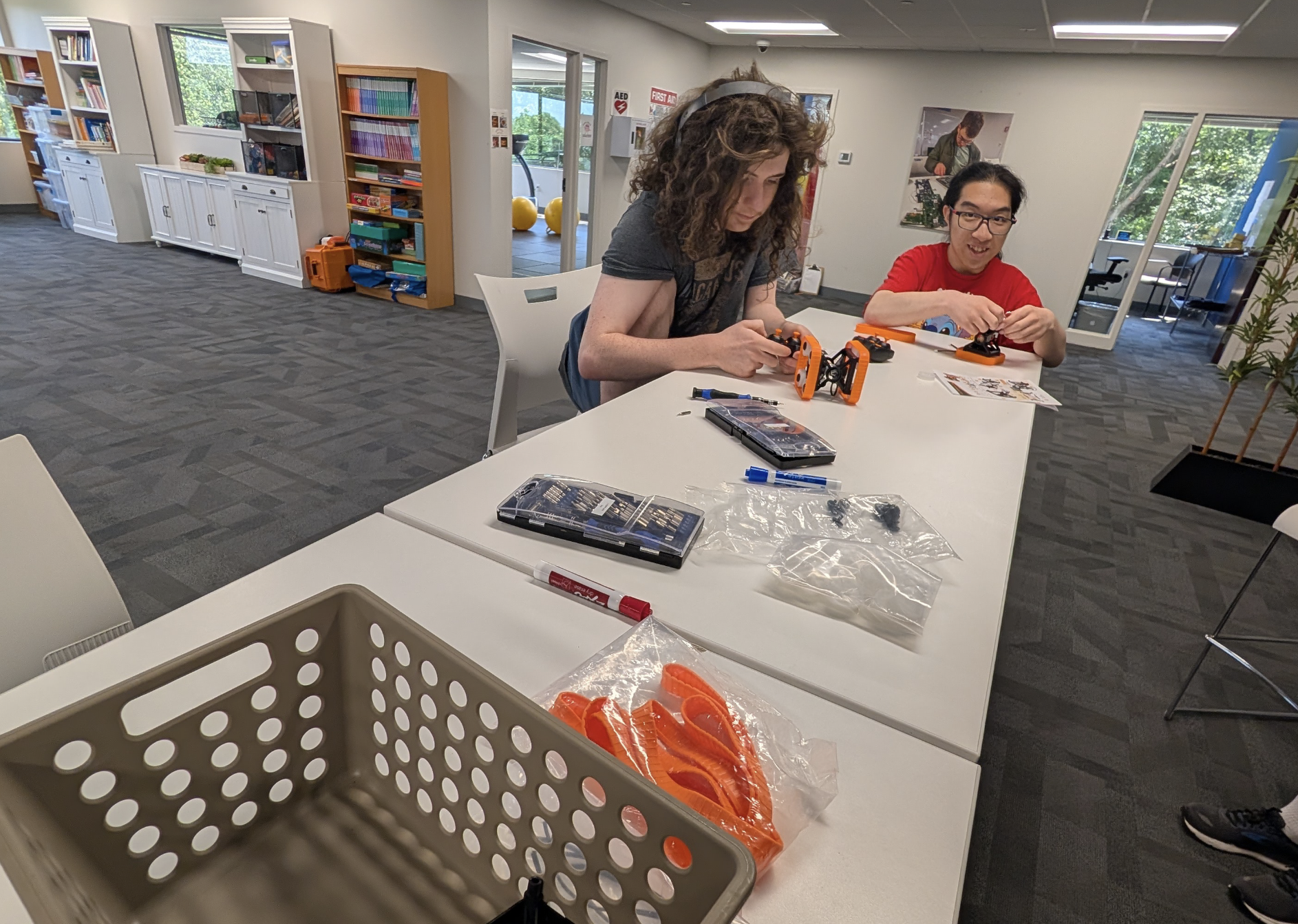 Two people working on assembling robots at a white table in a modern, well-lit room. Items like tools, parts, and instruction sheets are scattered on the table. The room has bookshelves, windows, and a door leading outside, with a few chairs and desk