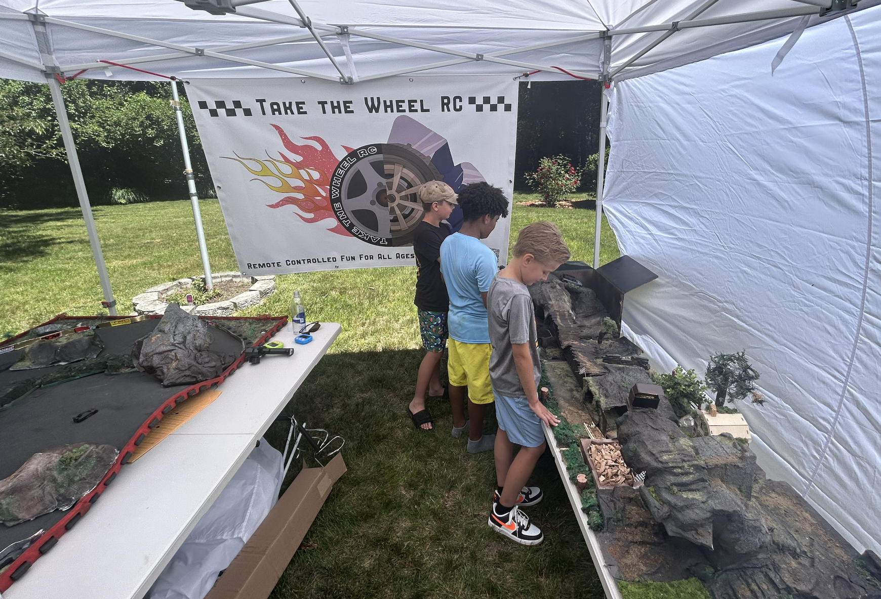 Four boys are standing and observing a model of a rocky landscape under a white canopy tent at an outdoor event. A banner behind them reads 'Take the Wheel RC' and 'Remote Controlled Fun for All Ages'.