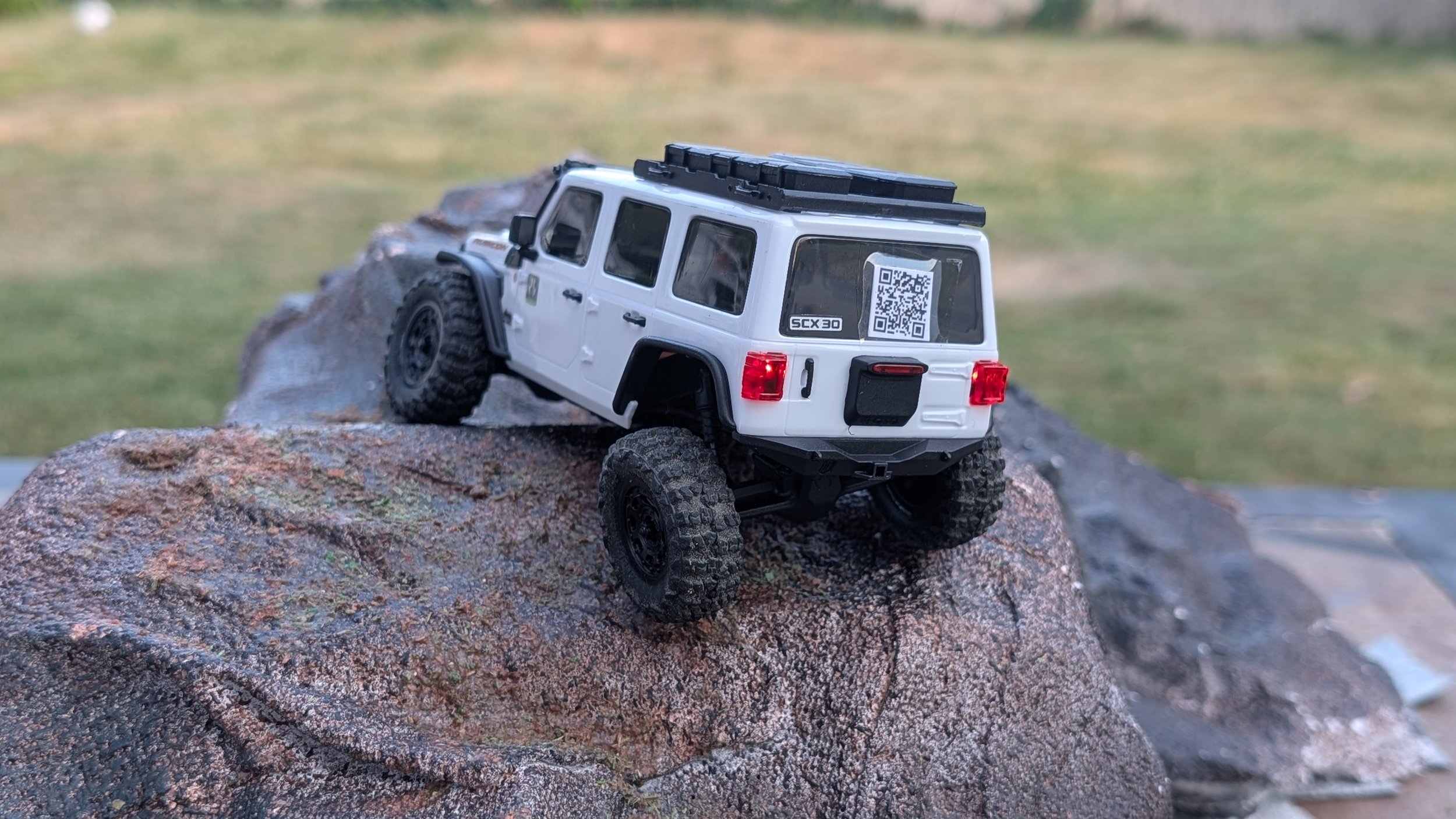 A remote-controlled model of a white off-road vehicle with large tires, positioned on a large rock outdoors with grass in the background.