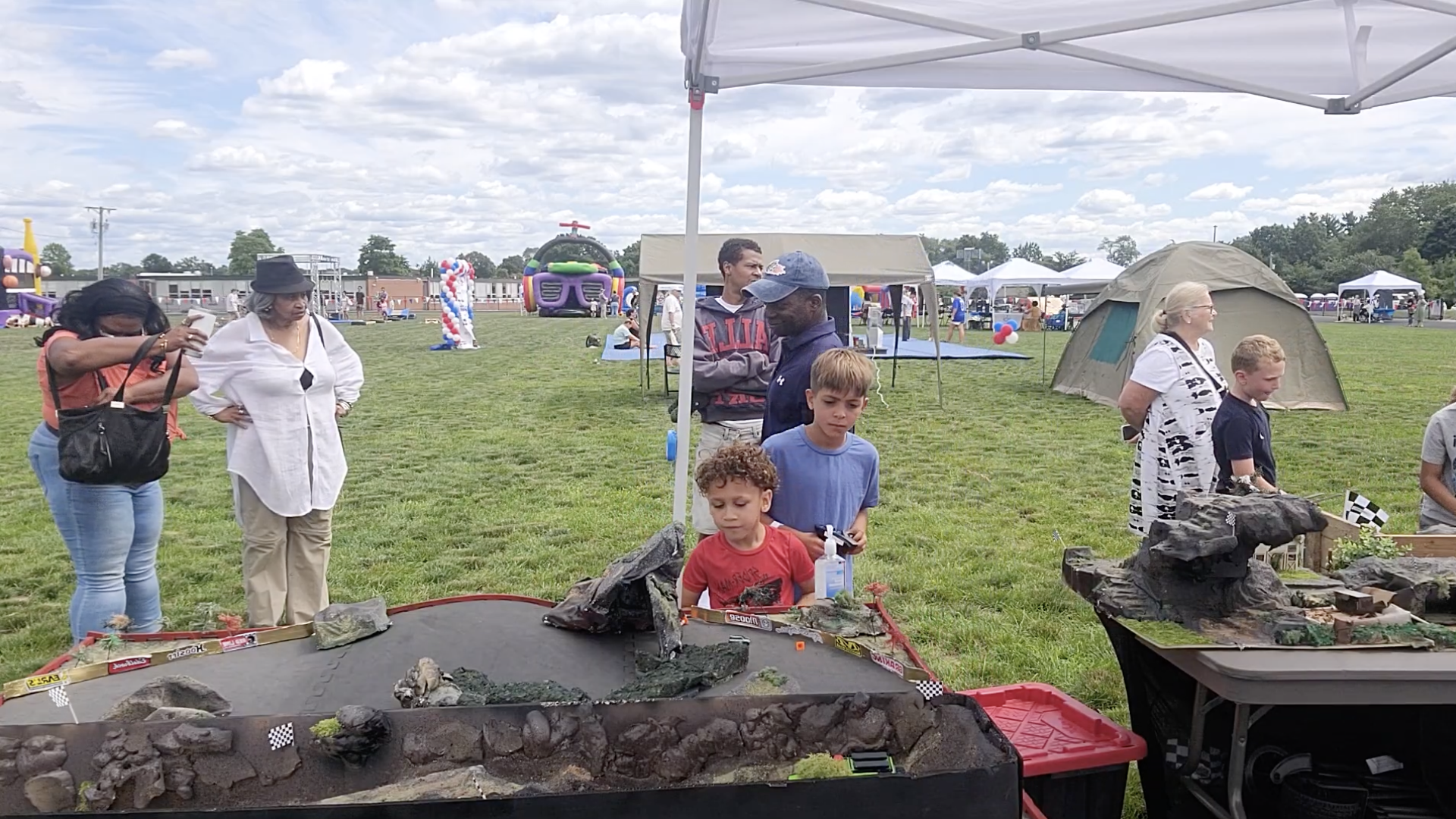 People gathered at Fredrick Flips Fair in Stoughton, with tents and bounce houses, looking at a miniature racing track with rocks and small models, on a grassy field under a partly cloudy sky.