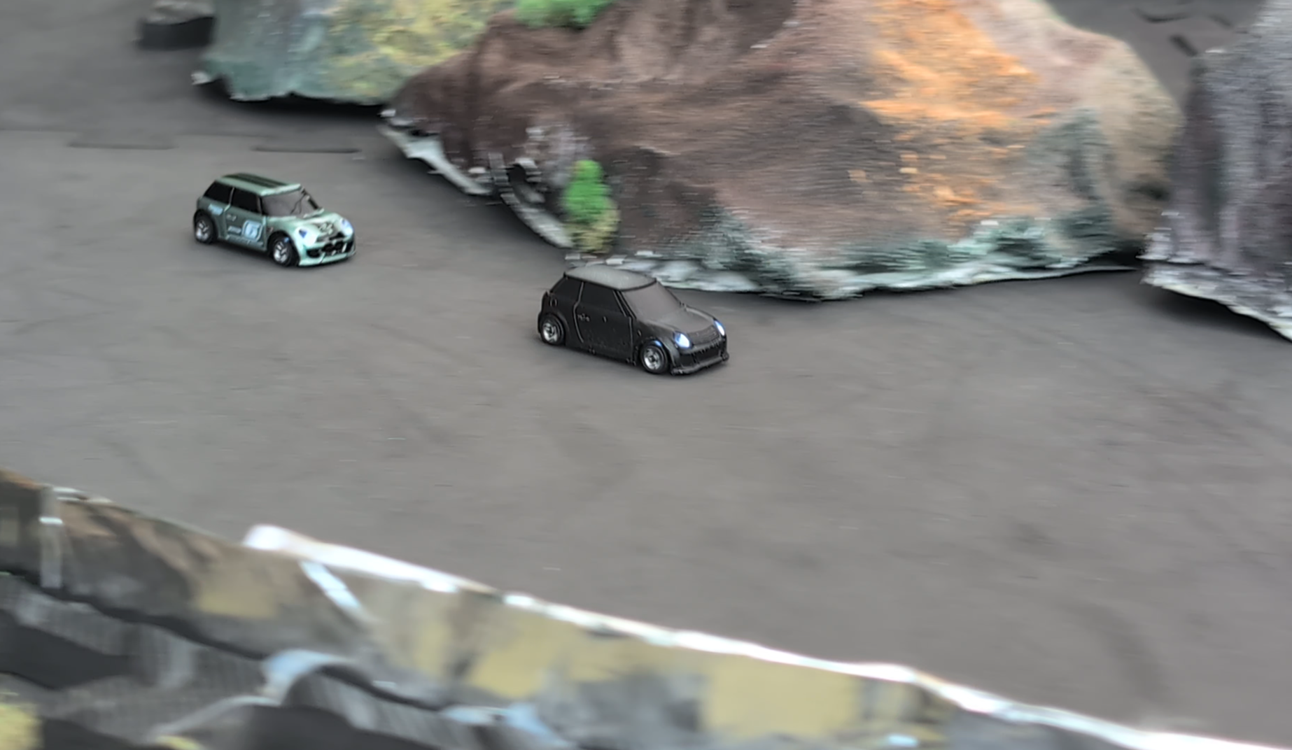 Two RC Mini Coopers, one matte black and one green with black roof, on a custom built race track with rocks and terrain in the background.