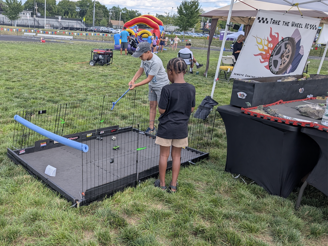 One child retrieves a stuck car in a small enclosed area at an outdoor event, with other people and a bounce house in the background.