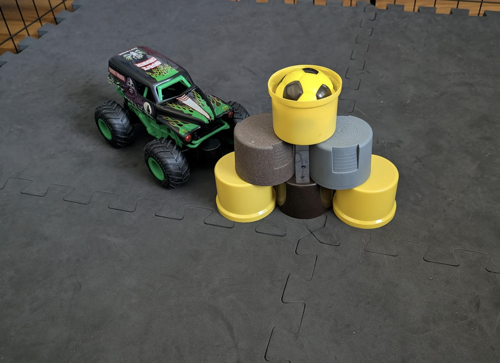 A toy monster truck parked next to a stack of plastic cylinders topped with a small yellow bucket containing a black and yellow soccer ball.